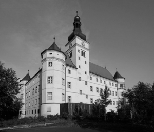 The black and white picture shows Schloss Hartheim, a white castle with four towers, surrounded by trees. 